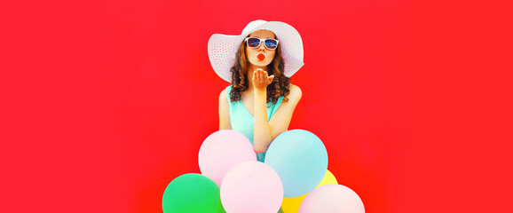Portrait of beautiful young woman with bunch of colorful balloons blowing her lips sends air kiss wearing summer straw hat on red background