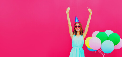 Naklejka na ściany i meble Happy cheerful smiling young woman in birthday hat with colorful balloons on pink background, blank copy space for advertising text