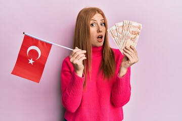 Young irish woman holding turkey flag and liras banknotes in shock face, looking skeptical and...