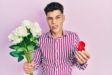 Young hispanic man holding bouquet of flowers and wedding ring clueless and confused expression....