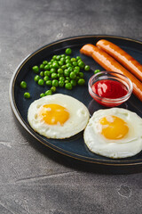 Classic breakfast of fried eggs, sausages and green peas on dish on gray background