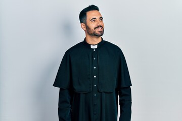 Handsome hispanic man with beard wearing catholic priest robe looking away to side with smile on...