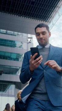 Caucasian Businessman in a Suit is Using a Smartphone on Dark Street in the Evening. Other Office People Walk Past. Video Footage with Vertical Screen Orientation 9:16.