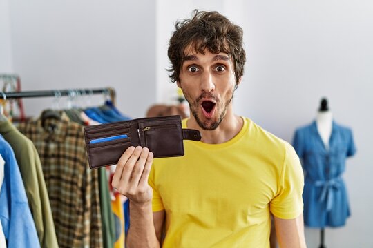 Young hispanic man holding leather wallet at retail shop scared and amazed with open mouth for surprise, disbelief face