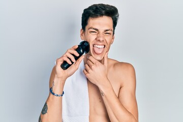 Young hispanic man shaving with electric razor machine sticking tongue out happy with funny expression.