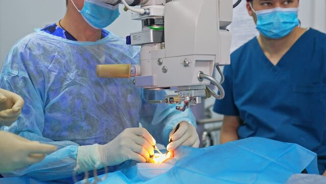 Male surgeon moves slowly with tools holding them in both hands. Chief doctor looks into huge microscope to conduct operation.