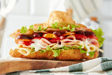 Croissant sandwich with cheese, ham, lettuce, tomatoes, olives and delicious sauce on wooden board.