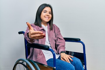 Young brunette woman sitting on wheelchair smiling friendly offering handshake as greeting and...
