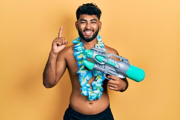 Arab man with beard wearing swimsuit and hawaiian lei holding watergun smiling with an idea or...