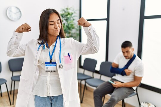 Young asian doctor woman at waiting room with a man with a broken arm showing arms muscles smiling proud. fitness concept.