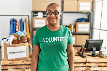 Mature hispanic woman wearing volunteer t shirt at donations stand looking positive and happy standing and smiling with a confident smile showing teeth