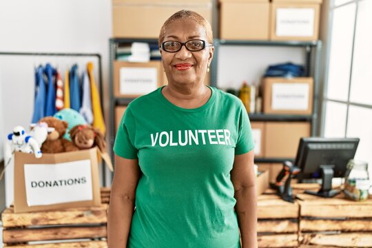 Mature hispanic woman wearing volunteer t shirt at donations stand looking positive and happy standing and smiling with a confident smile showing teeth