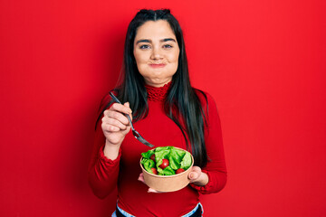 Young hispanic girl eating salad puffing cheeks with funny face. mouth inflated with air, catching...