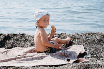 A boy child in Panama is sitting on the seashore, sunbathing and eating ice cream.