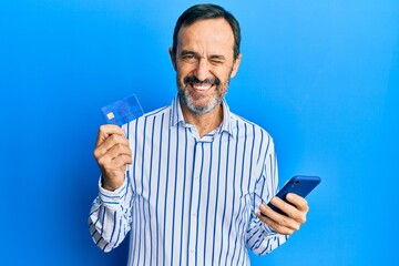 Middle age hispanic man using smartphone and holding credit card winking looking at the camera with...