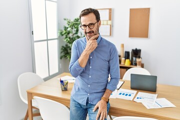 Middle age hispanic man with beard wearing business clothes at the office looking confident at the camera with smile with crossed arms and hand raised on chin. thinking positive.