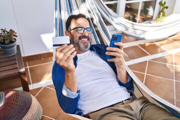 Middle age man using smartphone and credit card lying on hammock at terrace home