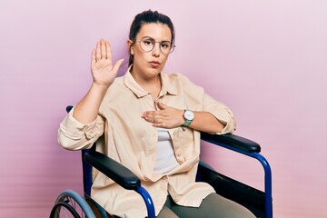Young hispanic woman sitting on wheelchair swearing with hand on chest and open palm, making a...