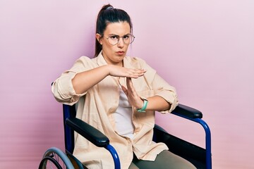 Young hispanic woman sitting on wheelchair doing time out gesture with hands, frustrated and...