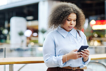 Waist up portrait of black young woman using smartphone in shopping mall wearing smart casual outfit, copy space