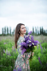 Beautiful girl with a bouquet of wild flowers. A girl in dress with a bouquet of lupine stands in flower field. Summertime. Blooming Lupine field. Summer vacation. Nature concept. space for text	
