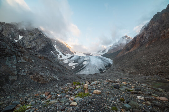 Scenic mountain landscape with large glacier in low clouds in sunrise colors. Colorful view to glacier tongue in morning sunlight in low clouds. Atmospheric high mountain scenery at early morning.