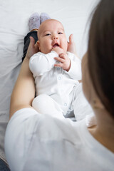 young woman holds an asian baby who looks at her mother and smiles while sitting on the bed at home. mom and baby,smile,motherhood, mother and baby at home. selective focus