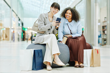 Full length portrait of two smiling young women using smartphone in shopping mall while sitting in rest area with bags