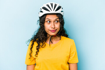 Young hispanic woman wearing a helmet bike isolated on blue background confused, feels doubtful and unsure.