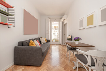 Living room with wooden and metal table for two people, wooden chairs, gray sofa bed with lots of cushions and colored shelves, balcony with white wooden shutters