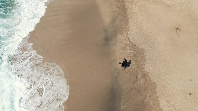 A gold digger walks along a sandy beach with a hand-held metal detector along the waves rolling ashore. Aerial filming from a drone