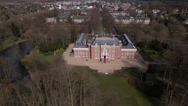 Sideways aerial pan around Slot Zeist castle with the moated manor surrounded by green park and urban landscape in the background. Dutch stately venue seen from above.