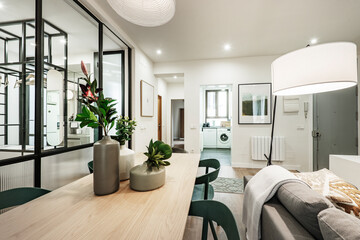 Living room with wooden dining table with vases and flowers, hanging lamps, sofa with blankets and entrance to a kitchen