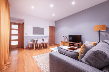 Living room with a white dining table, wooden furniture, a three-seater sofa, mustard-colored curtains and a wooden floor, and a tv on the sideboard
