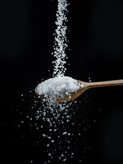 Edible salt crystals falling down into the wooden spoon at black background.