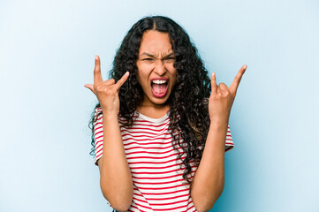 Young hispanic woman isolated on blue background showing a horns gesture as a revolution concept.