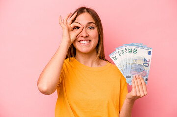 Young caucasian woman holding banknotes isolated on pink background excited keeping ok gesture on eye.
