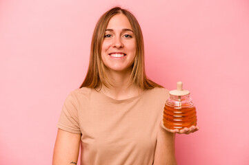 Young caucasian woman holding honey isolated on pink background laughing and having fun.