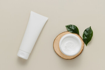 White cosmetic tube and bottle of cream on wooden stand with plant leaves top view, product design...