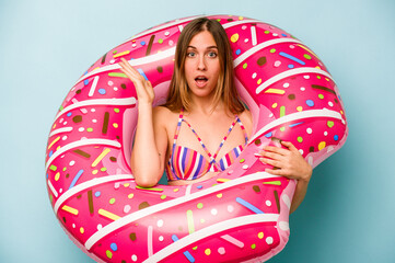 Young caucasian woman holding air mattress isolated on blue background surprised and shocked.