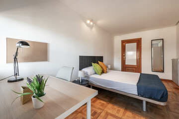 Bedroom with double bed with blue bed blanket, desk with plant and lamp, oak parquet flooring and beveled mirror hanging on the wall