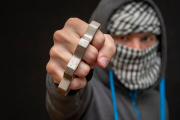 Brass knuckles in the hand of a man with a scarf covering his face on a dark background, selective focus.  Concept: street fights of hooligans, attacks on citizens, urban crime.