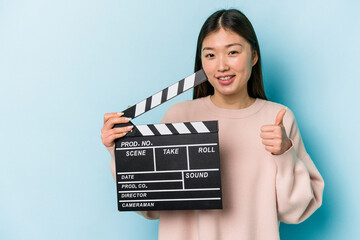 Young asian woman holding clapperboard isolated on blue background smiling and raising thumb up