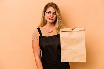 Restaurant waiter caucasian woman holding a take away bag isolated on beige background confused, feels doubtful and unsure.