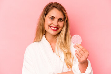 Young caucasian woman holding facial sponge isolated on pink background laughing and having fun.