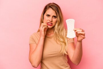 Young caucasian woman holding sanitary napkin isolated on pink background biting fingernails, nervous and very anxious.
