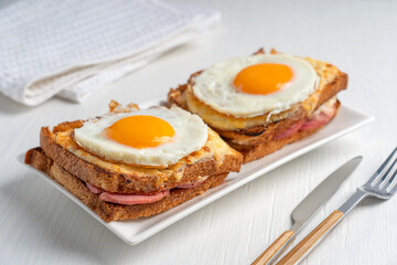 Homemade croque madame french hot sandwich made of toasted bread, ham and cheese with fried egg on top baked in oven served on plate on white wooden table with tableware for breakfast as quick snack