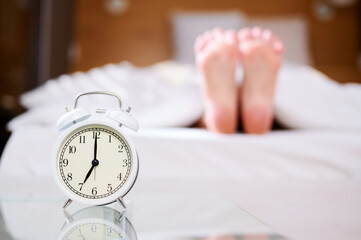 Sleeping woman legs in bed, early morning, alarm clock ringing. Selective focus. Laziness, lateness