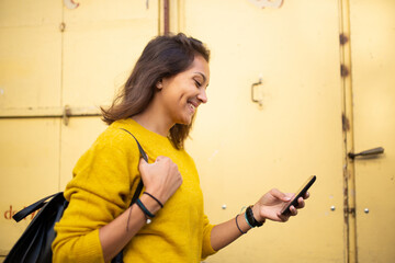 Side of smiling young woman looking at cellphone by yellow wall