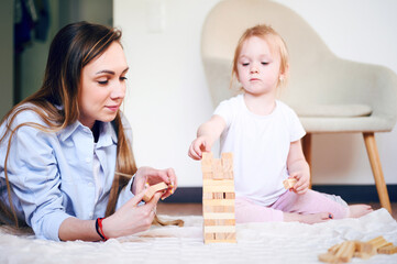Obraz na płótnie Canvas Young mother plays jenga with her cute preschool daughter on the floor at home. Happy family have fun at home